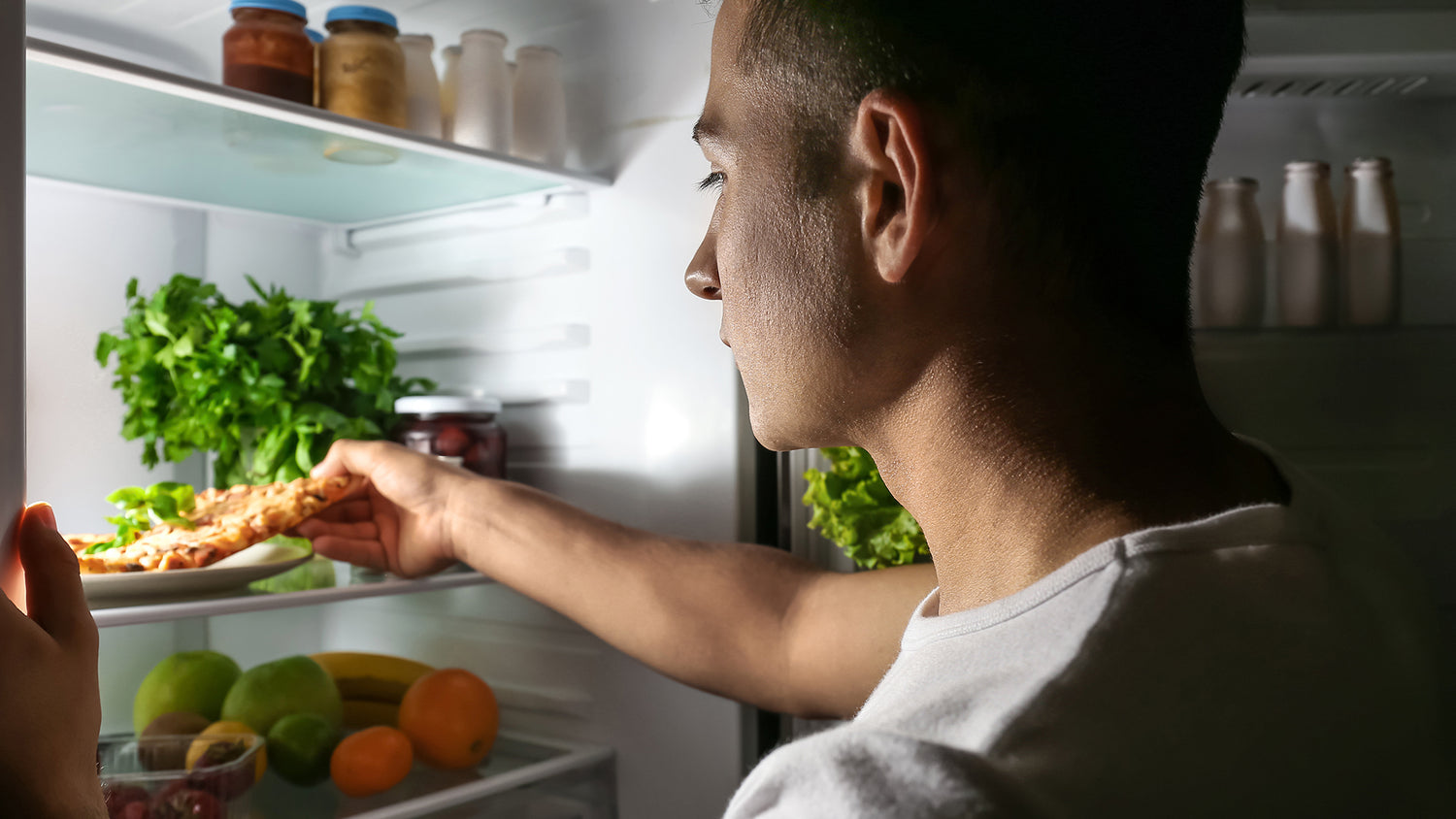 Side profile of man reaching into lit fridge for cold pizza slice at night, article 5 Eating Habits That Are Zapping Your Energy.