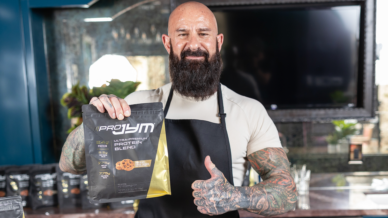 Smiling tattooed man in white shirt and black apron holds Pro JYM Chocolate Cookie Crunch pouch, Protein Powder For Fat Loss.