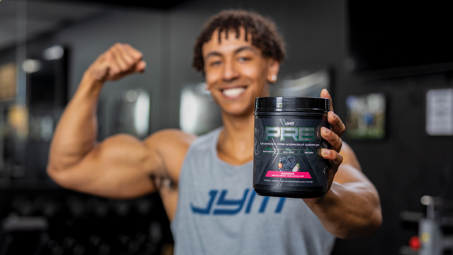 Grinning athlete in JYM tank flexes one arm and displays the DragonBerry Pre JYM X pre-workout tub label forward.