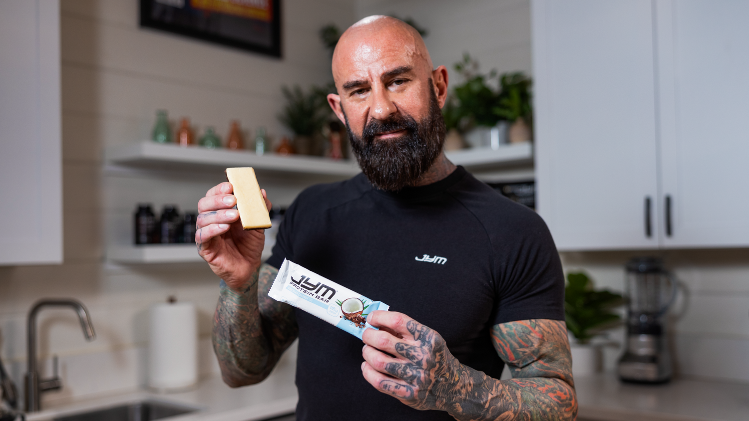 Bald bearded man in black JYM shirt holds piece of coconut JYM Protein Bar and blue wrapper in modern kitchen.