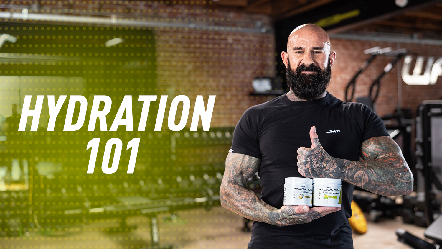 Hydration 101: bald, bearded man in black JYM shirt holds two Hydration JYM electrolyte powder tubs in a gym, thumbs up.