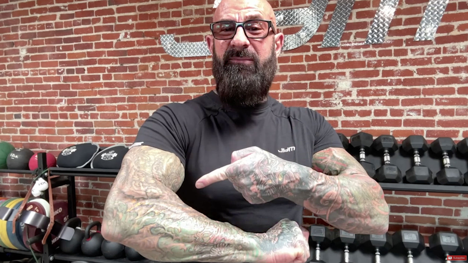 Jim Stoppani crossing tattooed forearms at chest in JYM gym, introducing three-step system for bigger forearms article.