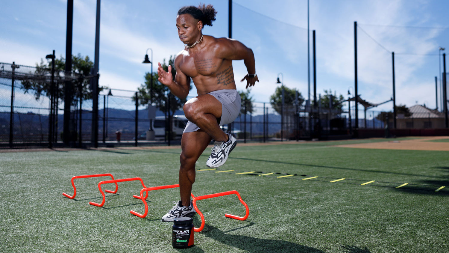 Muscular athlete leaping over red agility hurdles on sunlit turf field, Post JYM Carb container below — Better Brain Function with HIIT.