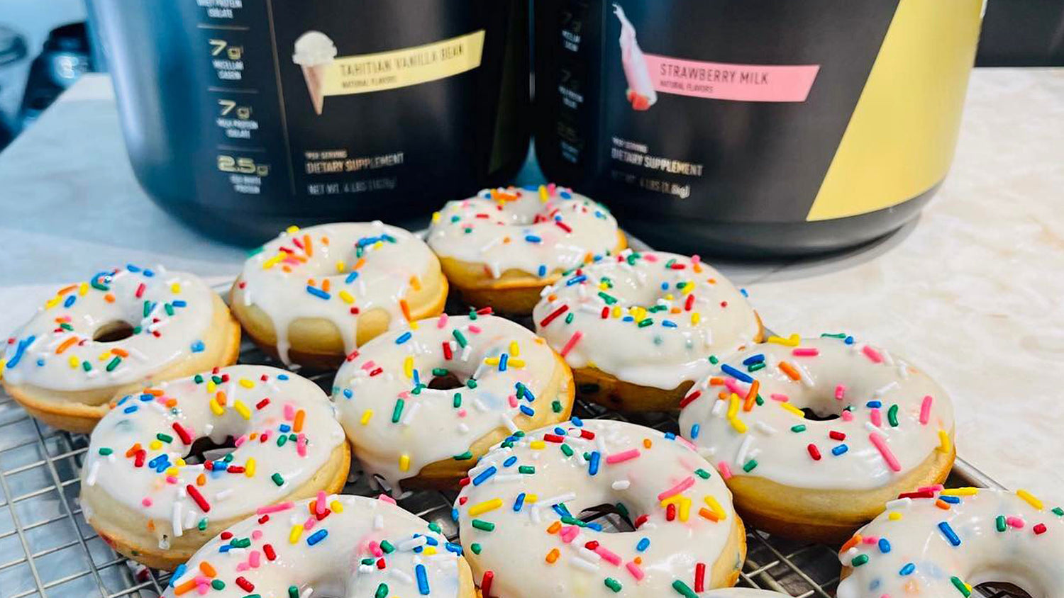 Glazed Birthday Cake Protein Donuts topped with rainbow sprinkles on a wire rack, with two Pro JYM protein tubs behind them.