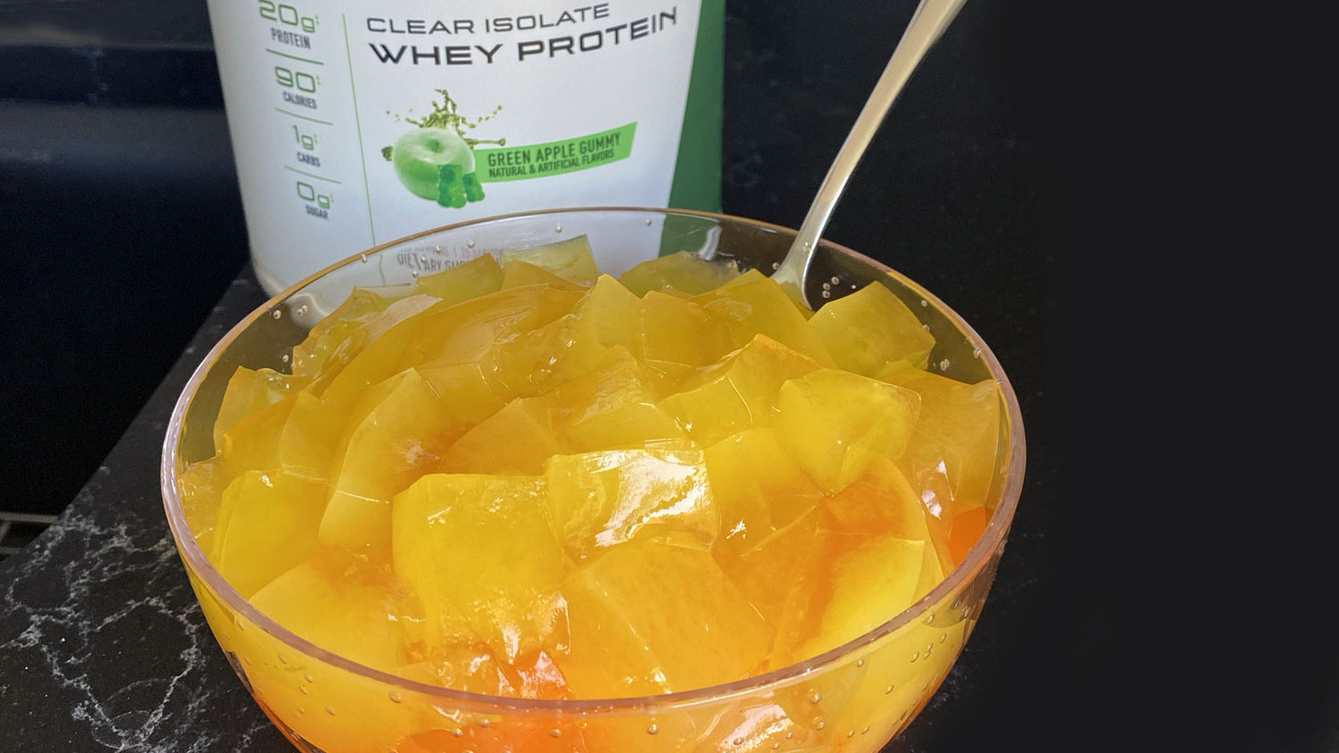 Glass bowl of orange Clear Protein Jello cubes with spoon, beside Green Apple Gummy Clear Isolate Whey Protein tub.