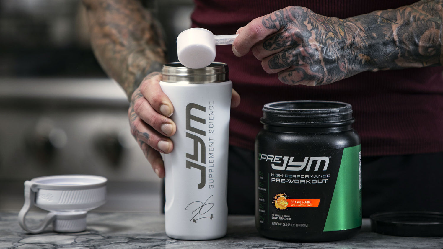 Tattooed arms holding JYM shaker as scoop of Pre JYM Orange Mango powder is added, tub beside on counter.