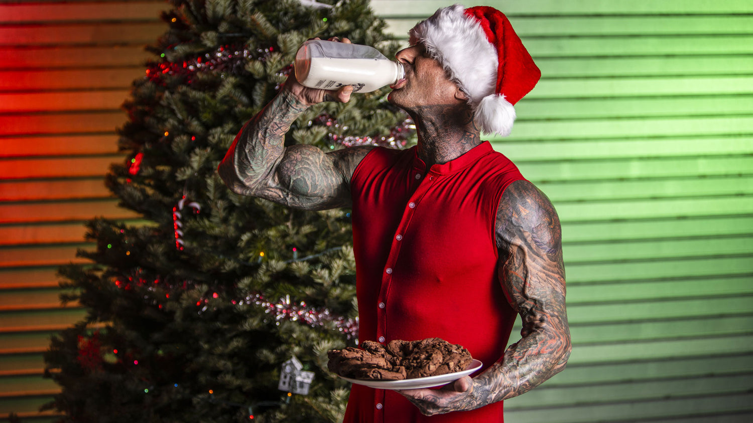 Tattooed athlete in Santa hat and red shirt drinking milk, cookies in hand, by a decorated tree—Holiday Tracking article.