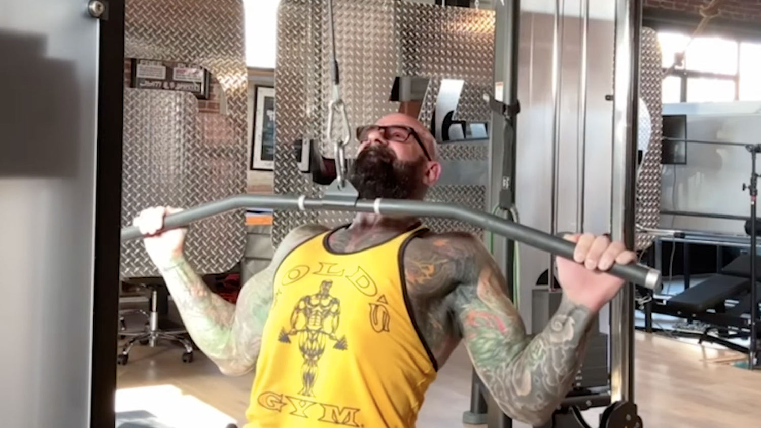 Tattooed Jim Stoppani leans back gripping a curved lat pulldown bar overhead, wearing a yellow Gold's Gym tank in a home gym.