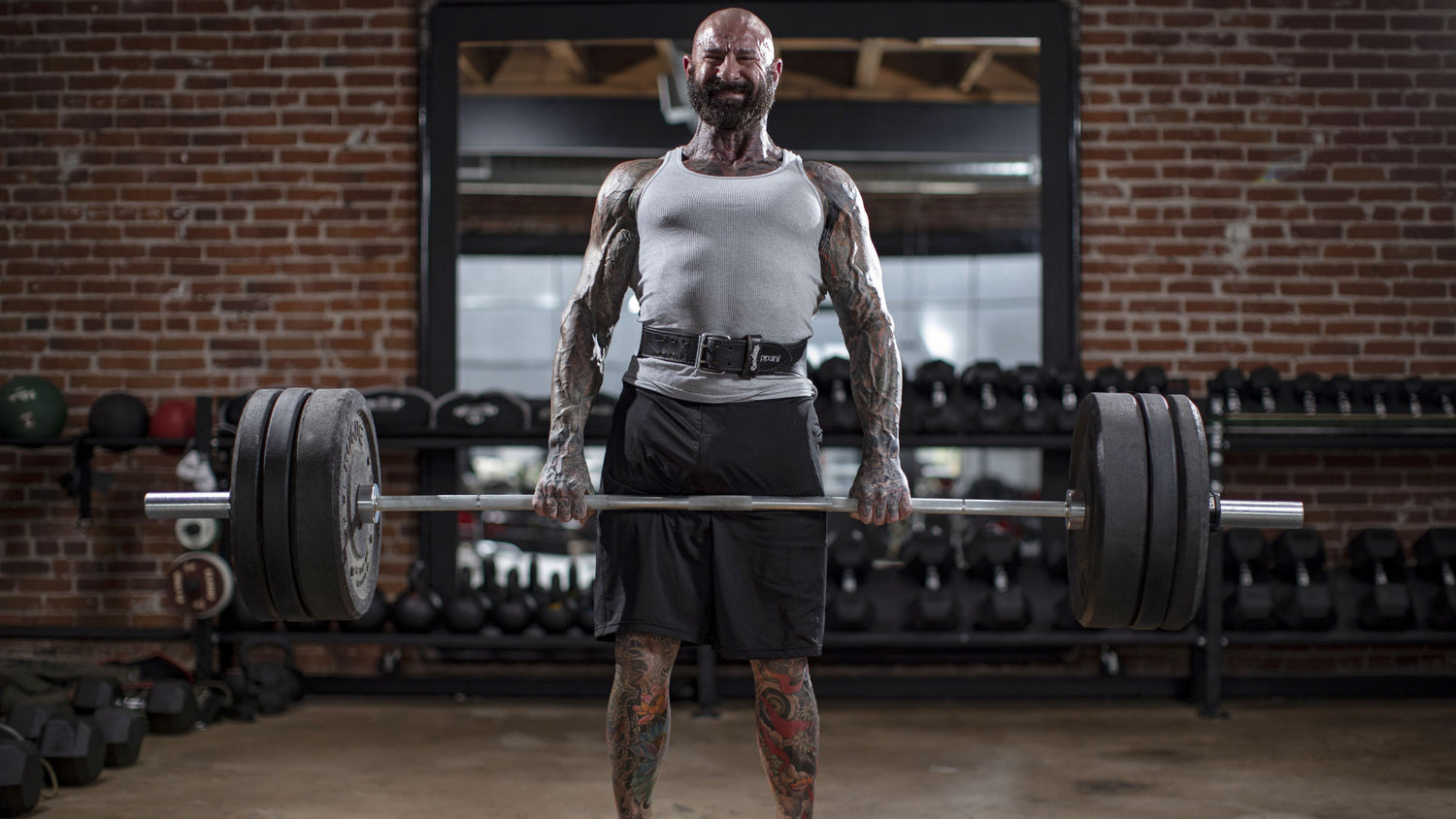 Tattooed bodybuilder locking out a loaded barbell deadlift in front of a mirror and dumbbell rack, Periodization article header.