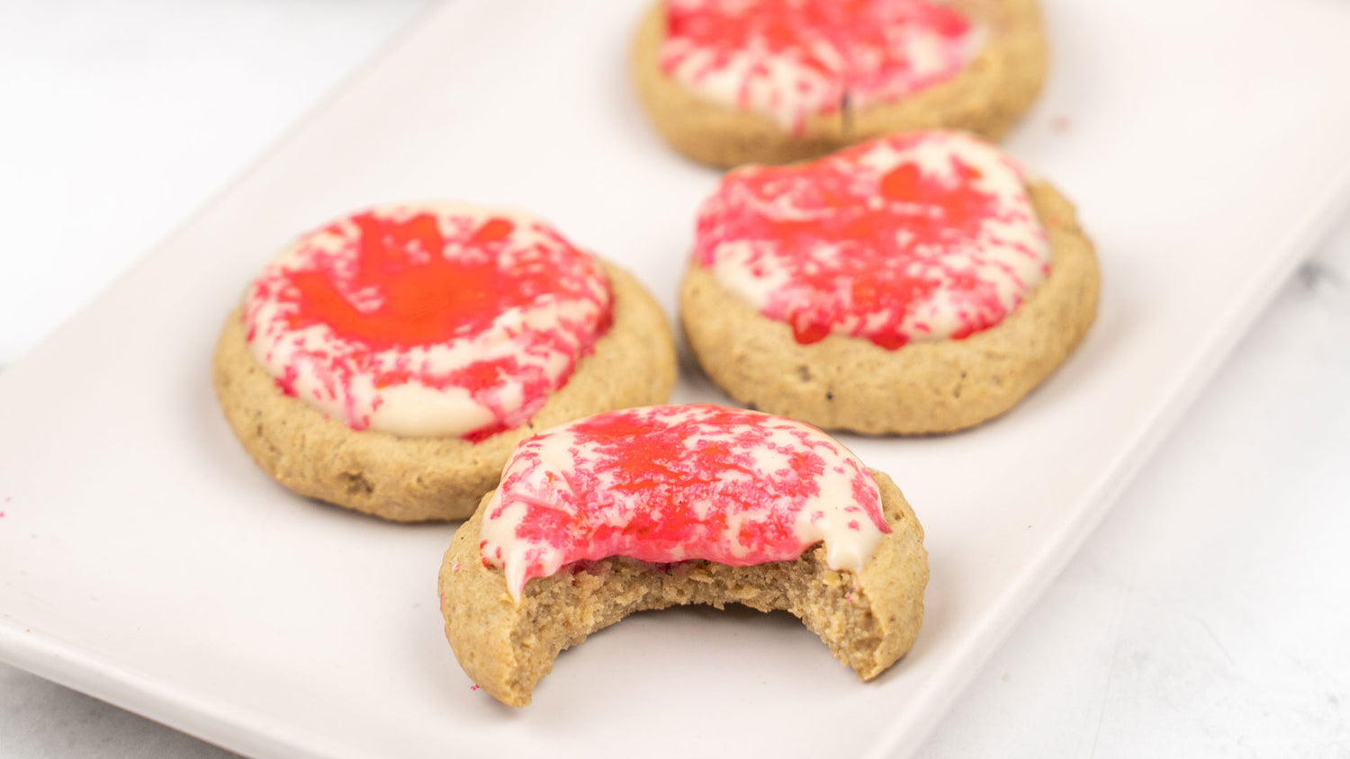 Four Frosted Gingerbread Protein Cookies with red and white swirled icing on a white tray, front cookie with a bite taken.