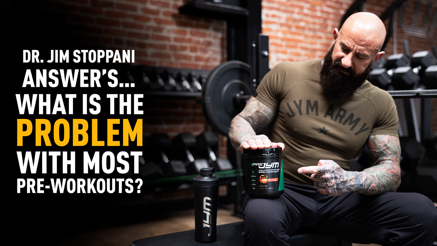 Dr. Jim Stoppani in olive JYM Army tee holds Pre JYM Cherry Limeade tub on gym bench with shaker and weight rack behind.