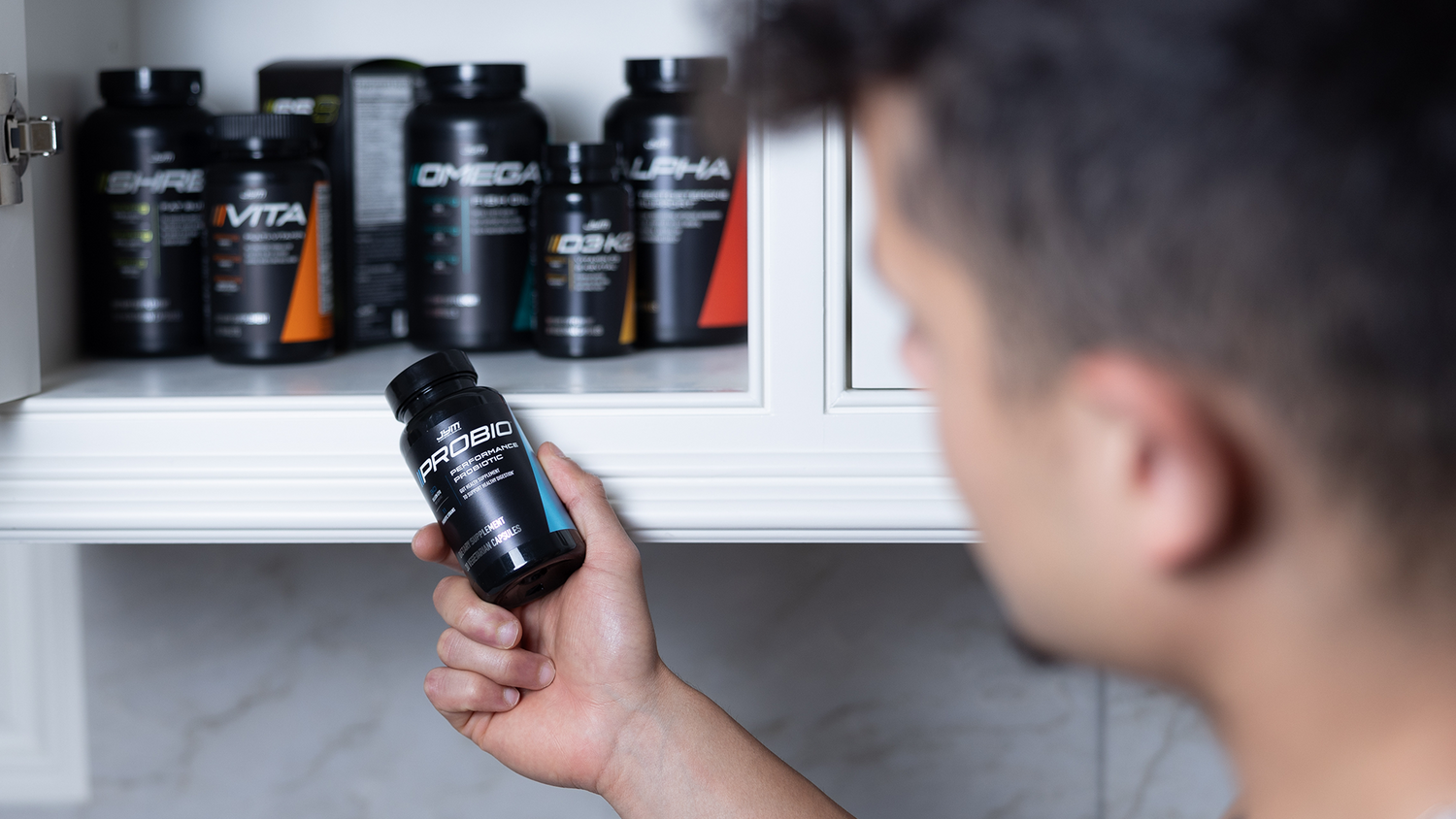 Close-up of hand gripping JYM Probio capsule bottle, with Shred, Vita, Omega, D3 K2, and Alpha JYM bottles on shelf behind.