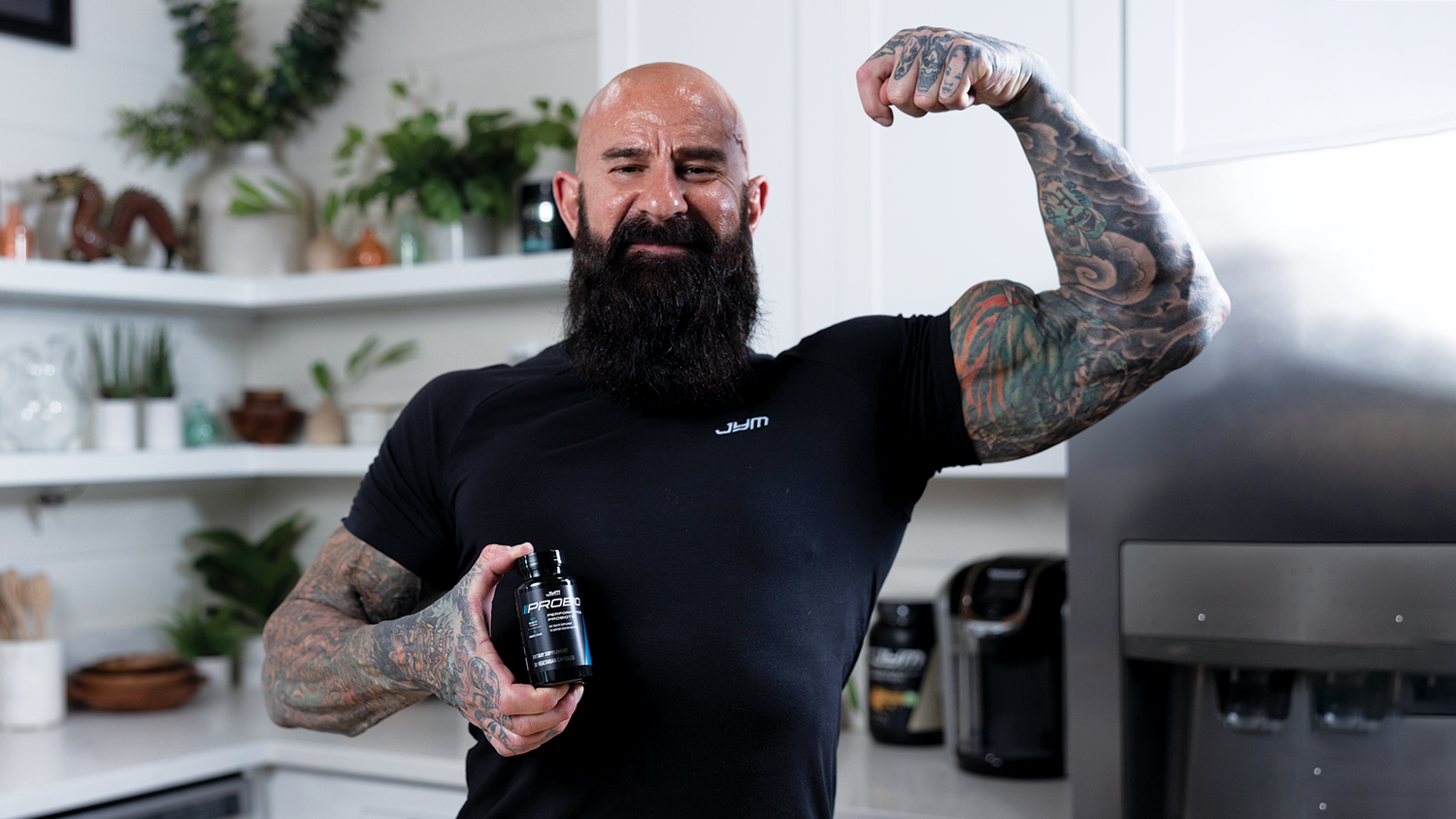 Muscular tattooed man with beard in black JYM shirt flexes arm, showing Pro JYM Probiotics bottle label in modern kitchen.