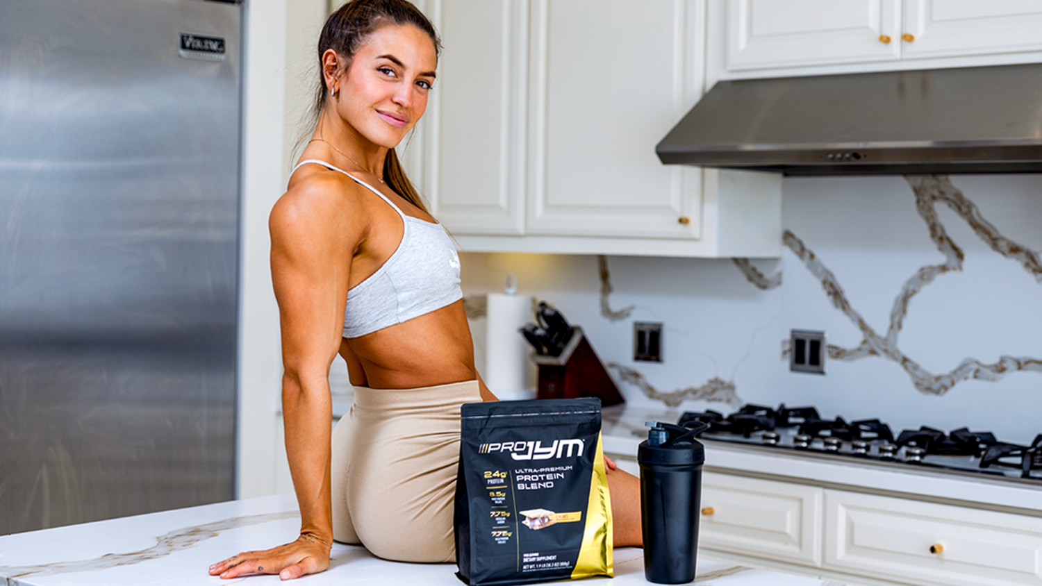 Woman in sports bra sitting on marble kitchen counter beside Pro JYM protein blend pouch and black shaker bottle.