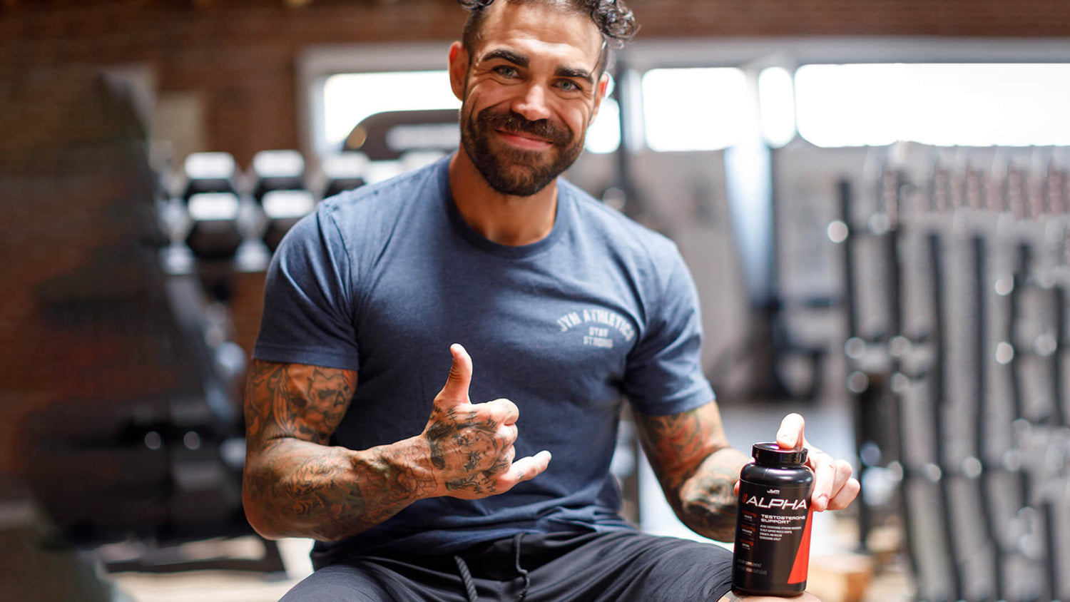 Smiling tattooed man in navy JYM Athletics tee holds black Alpha JYM testosterone support bottle, dumbbells blurred behind him.