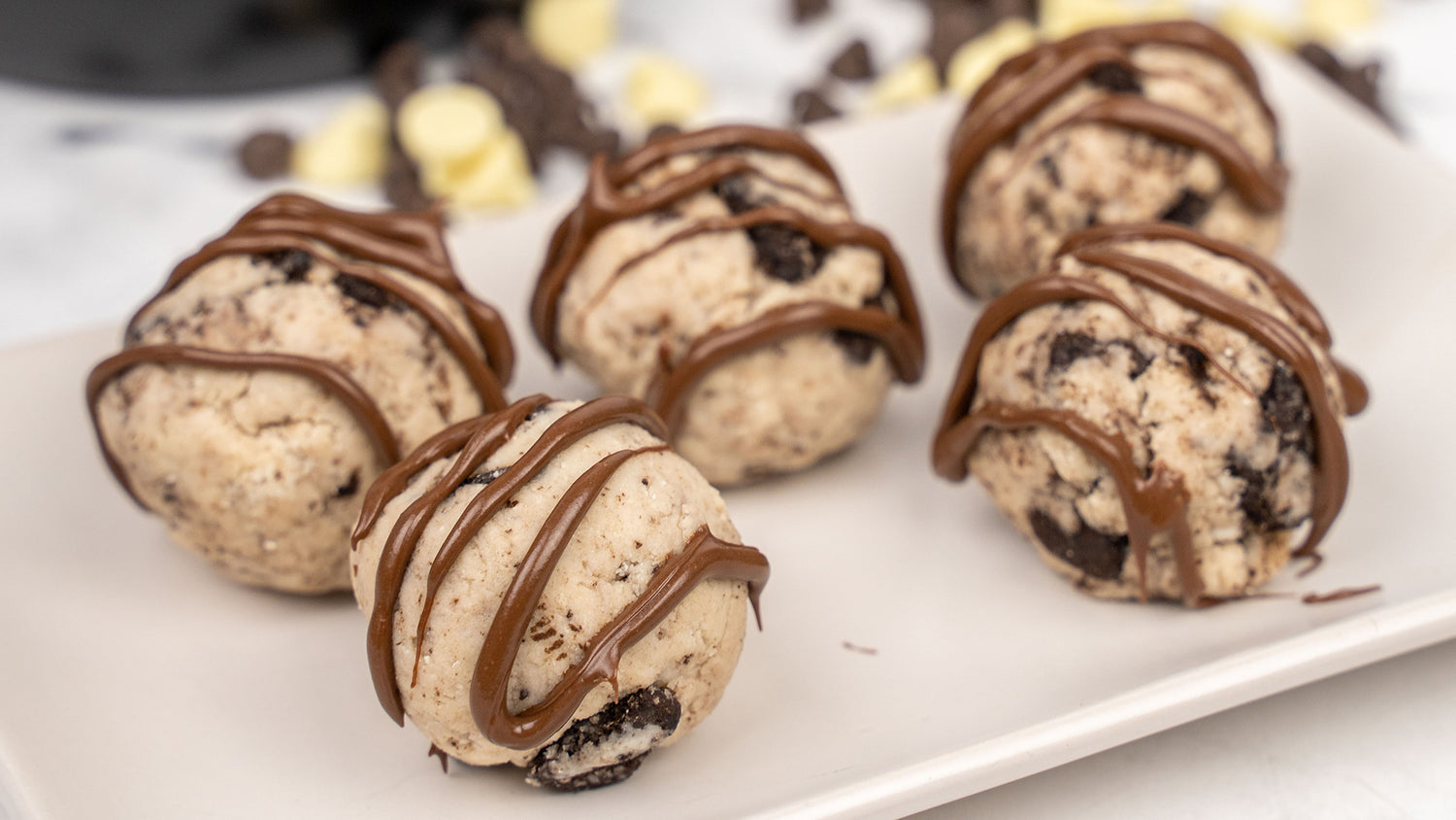 Five Cookies & Cream Protein Balls drizzled with chocolate on a white plate, with chocolate chips scattered behind.