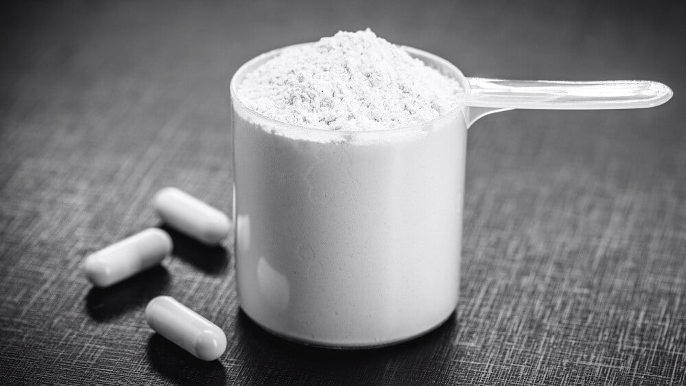Scoop heaped with white creatine powder beside three white capsules on a dark textured surface, from Creatine Myths Busted!