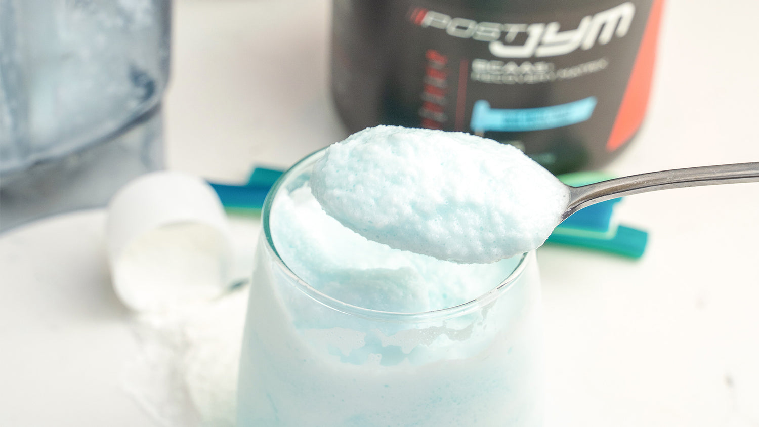 Close-up of blue slushie in a glass topped with foam, spoon scooping from above, Post JYM tub behind, for Blue Arctic Freeze recipe.