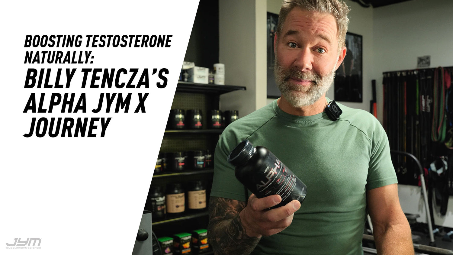 Tattooed man with gray beard holds Alpha JYM X bottle toward camera, JYM supplement shelves and barbell rack behind him.