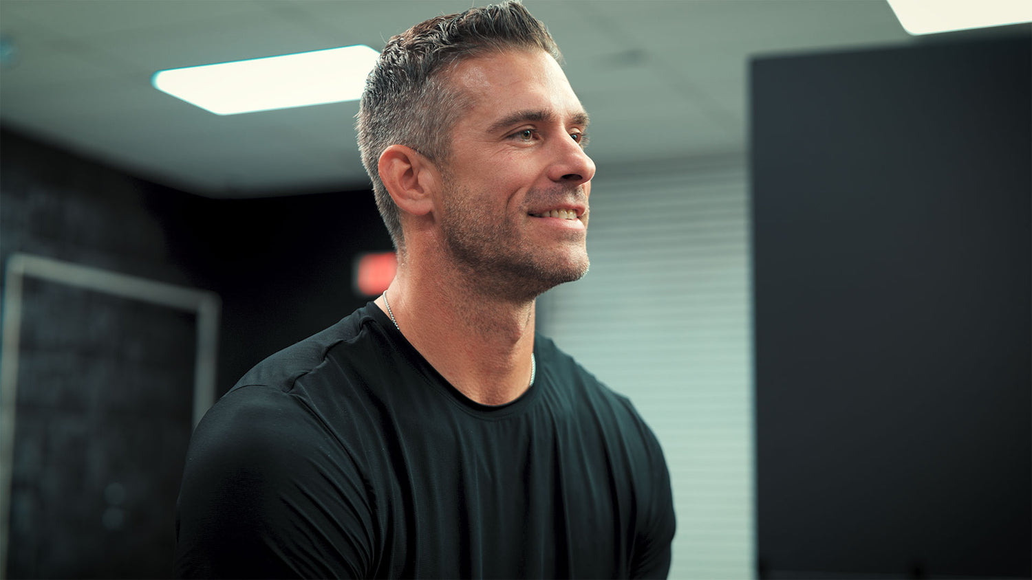 Jeff Wilk with short dark hair and stubble, grinning in a black tee under gym ceiling lights, Alpha JYM X testimonial.