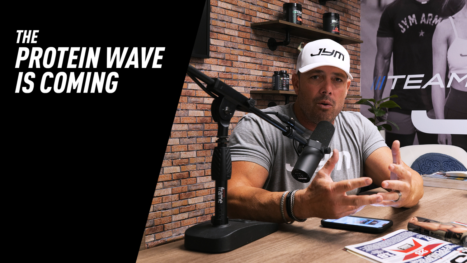 Podcast host in JYM hat gestures at Shure mic, JYM tubs on shelves behind, text reads The Protein Wave Is Coming.