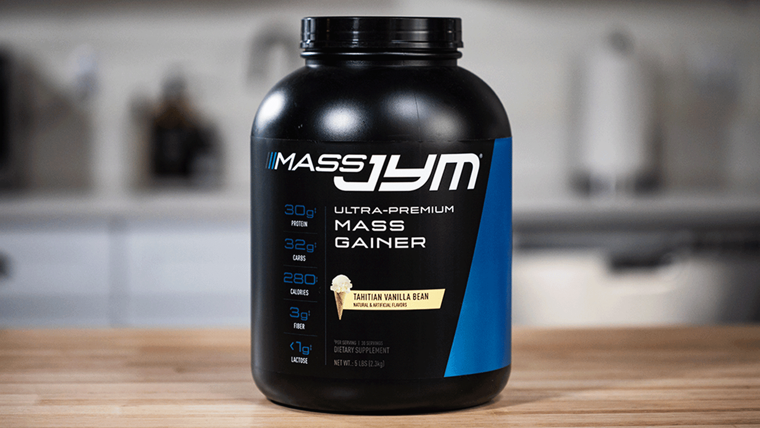 Black 5 lb Mass JYM Tahitian Vanilla Bean mass gainer tub on a wood countertop, nutrition stats listed down the front label.