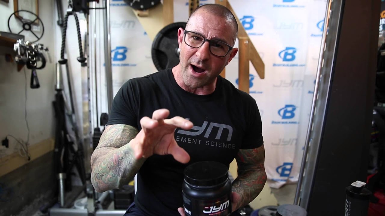 Dr. Jim Stoppani in a JYM t-shirt holding a black Pre JYM tub in a gym, discussing Caffeine-Free Pre JYM.