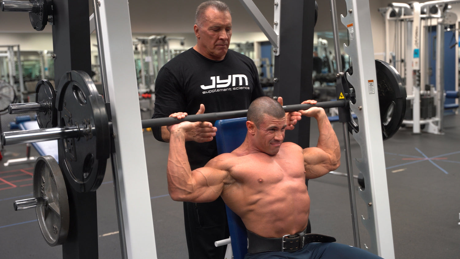 Milos Sarcev in black JYM tee spotting a shirtless bodybuilder mid-rep on Smith machine shoulder press with weight belt.