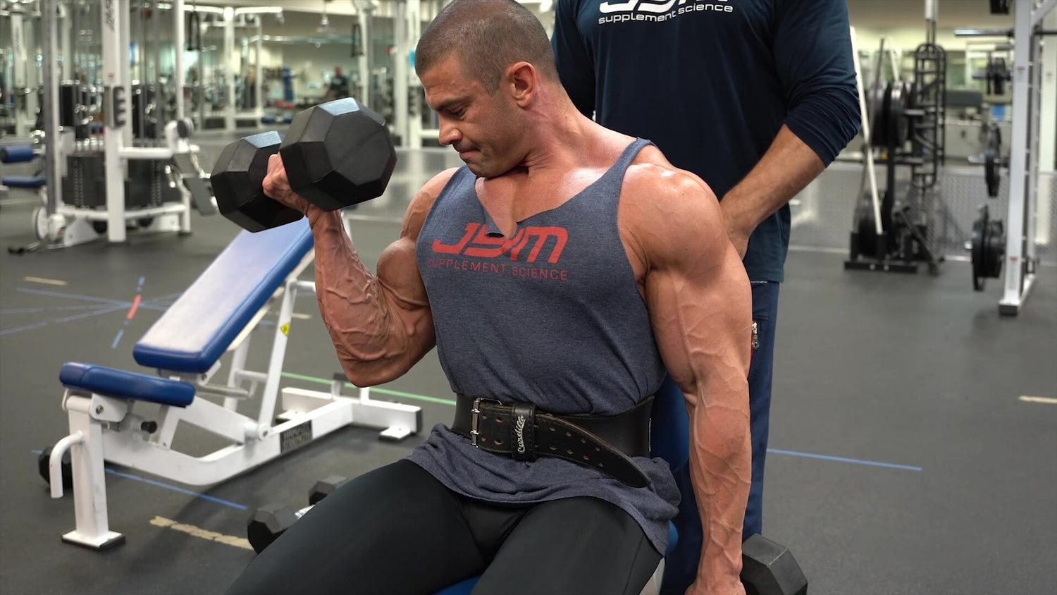 Muscular lifter in JYM Supplement Science tank performs seated dumbbell biceps curl with weight belt, focused on contraction.