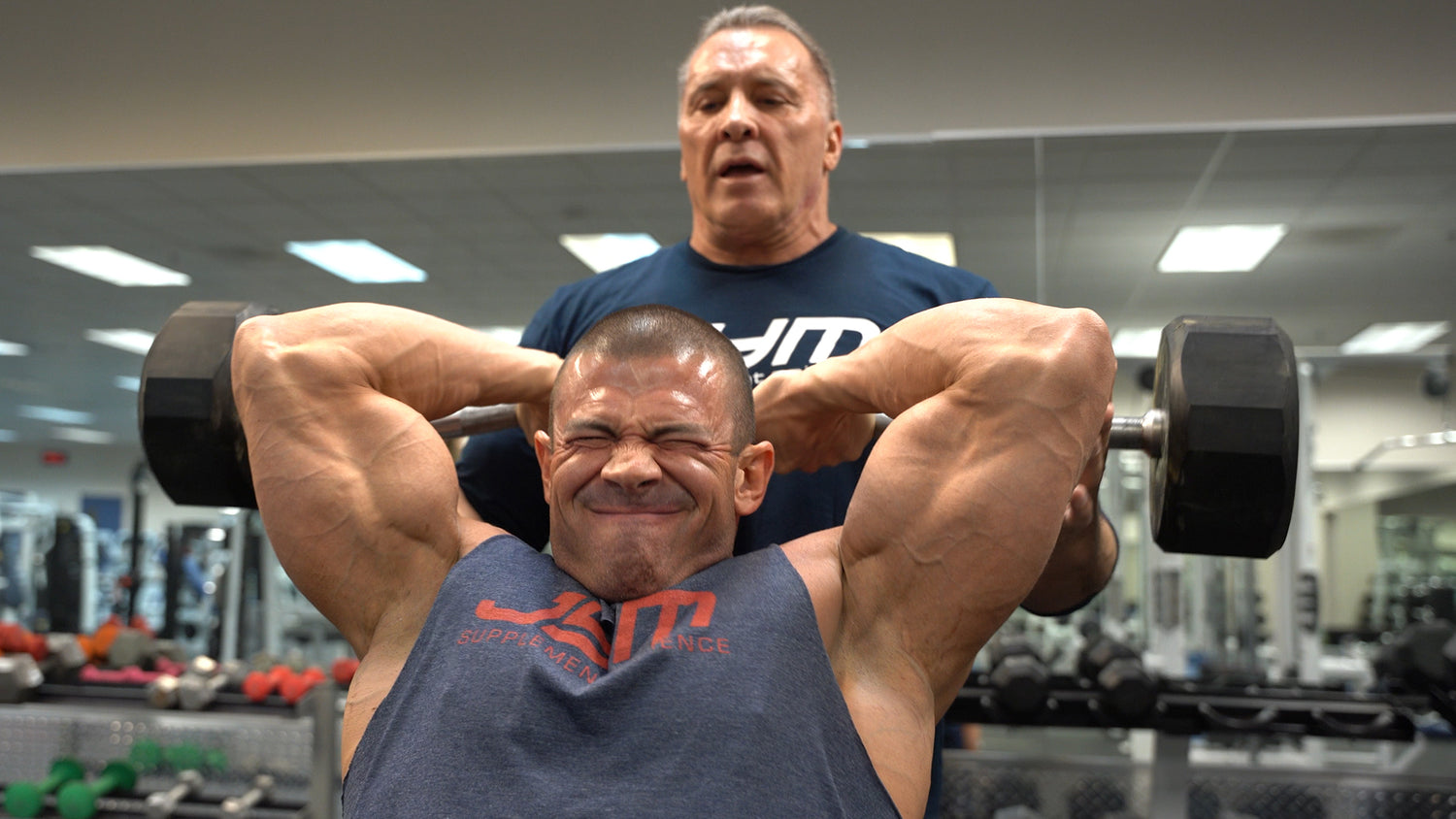 Milos Sarcev coaches a straining lifter in a JYM shirt lifting heavy dumbbells overhead for Monster Triceps giant sets.