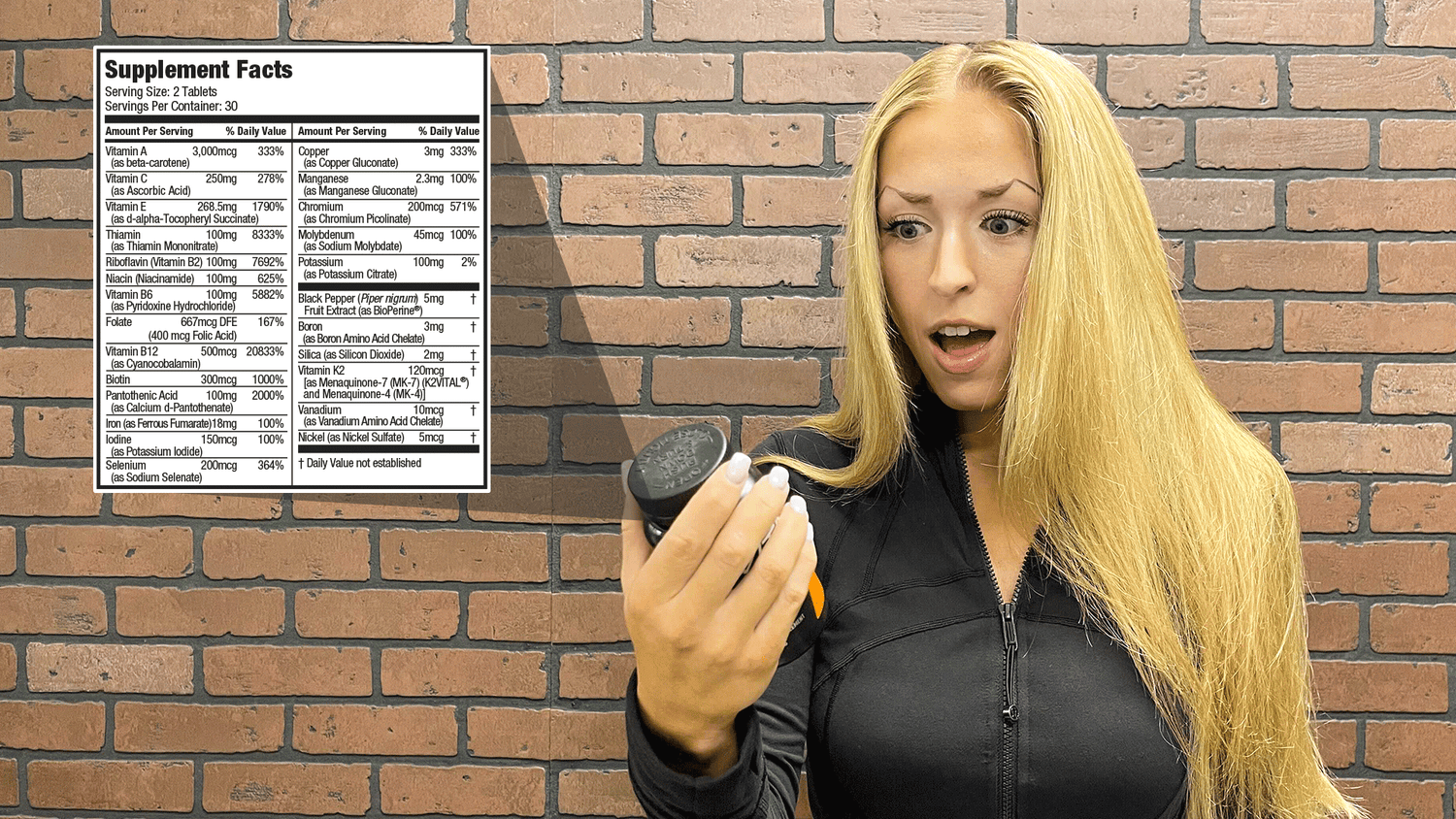 Shocked woman examining JYM supplement bottle in front of brick wall with detailed vitamin and mineral facts panel, for "Most Multivitamins Suck!".