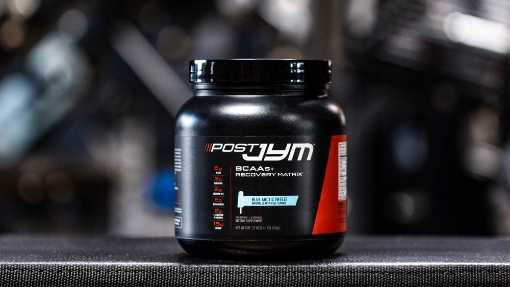 Black tub of Post JYM BCAAs+ Recovery Matrix in Blue Arctic Freeze flavor on a gym surface with blurred equipment behind.