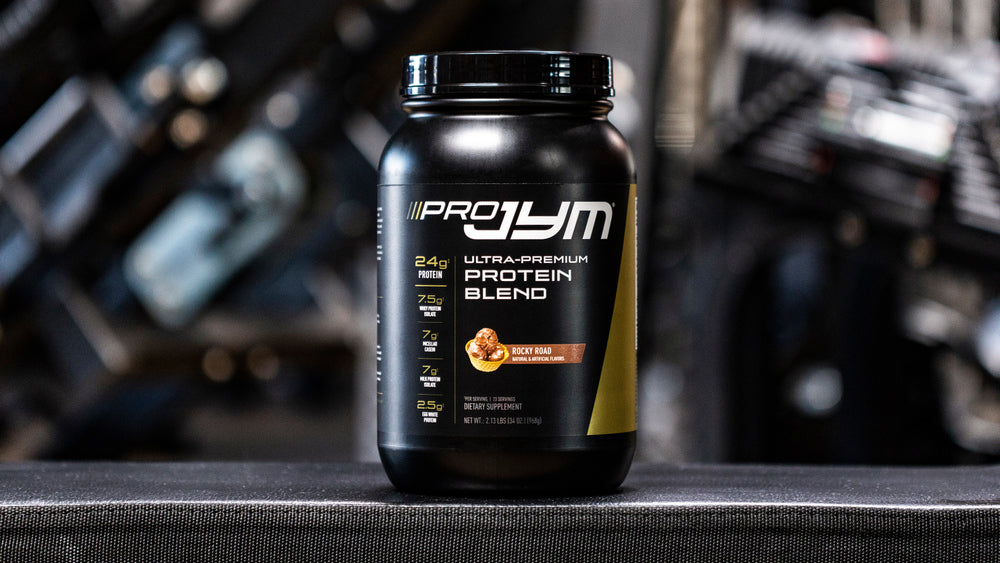 Pro JYM Rocky Road protein blend tub on gym bench with blurred weight rack background, front label showing 24g protein.