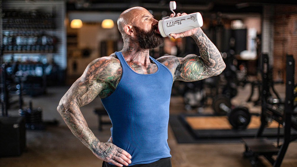 Tattooed bearded man in blue tank top drinking from a white JYM shaker bottle in a gym, illustrating Protein Powder Problems.