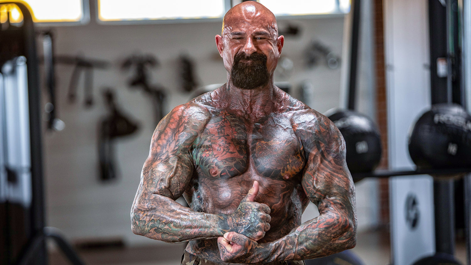 Shirtless Jim Stoppani facing camera, thumb up, tattooed arms and chest, blurred gym weights behind — Rest-Pause: A Proven Muscle-Building Method.