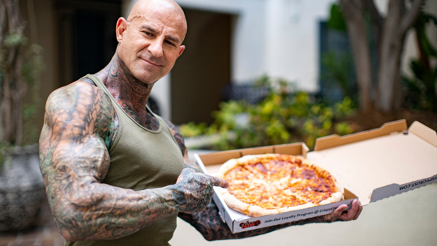 Jim Stoppani in green tank top displays open pizza box with cheese pizza in courtyard, for article on cheat meals.