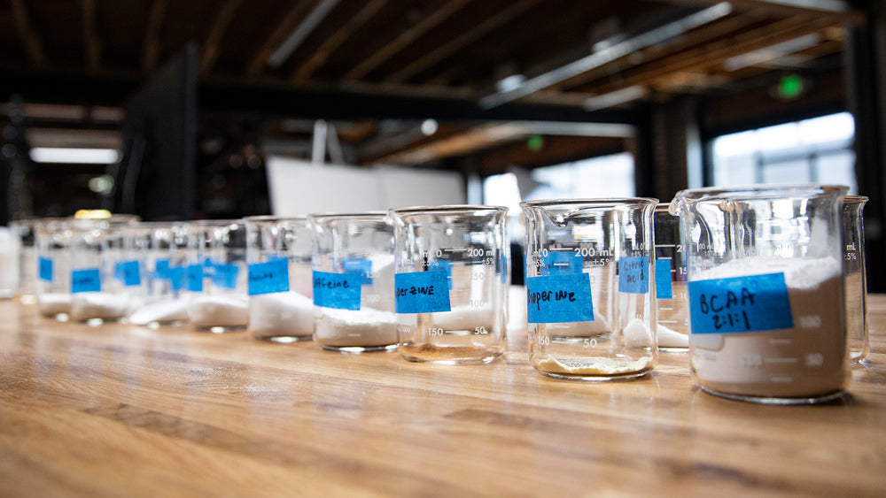 Row of 200 mL glass beakers with blue-taped labels holding white supplement powders on a wood bench for Supplement Guide.