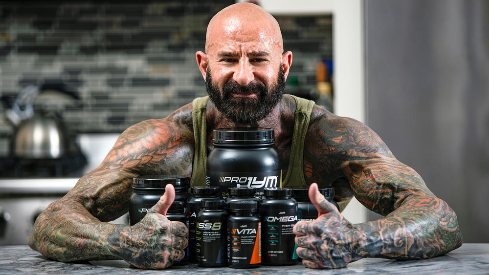 Muscular tattooed man with beard posing behind lineup of Pro JYM, SS8, Vita JYM and Omega JYM in kitchen, Supplements on Rest Days.