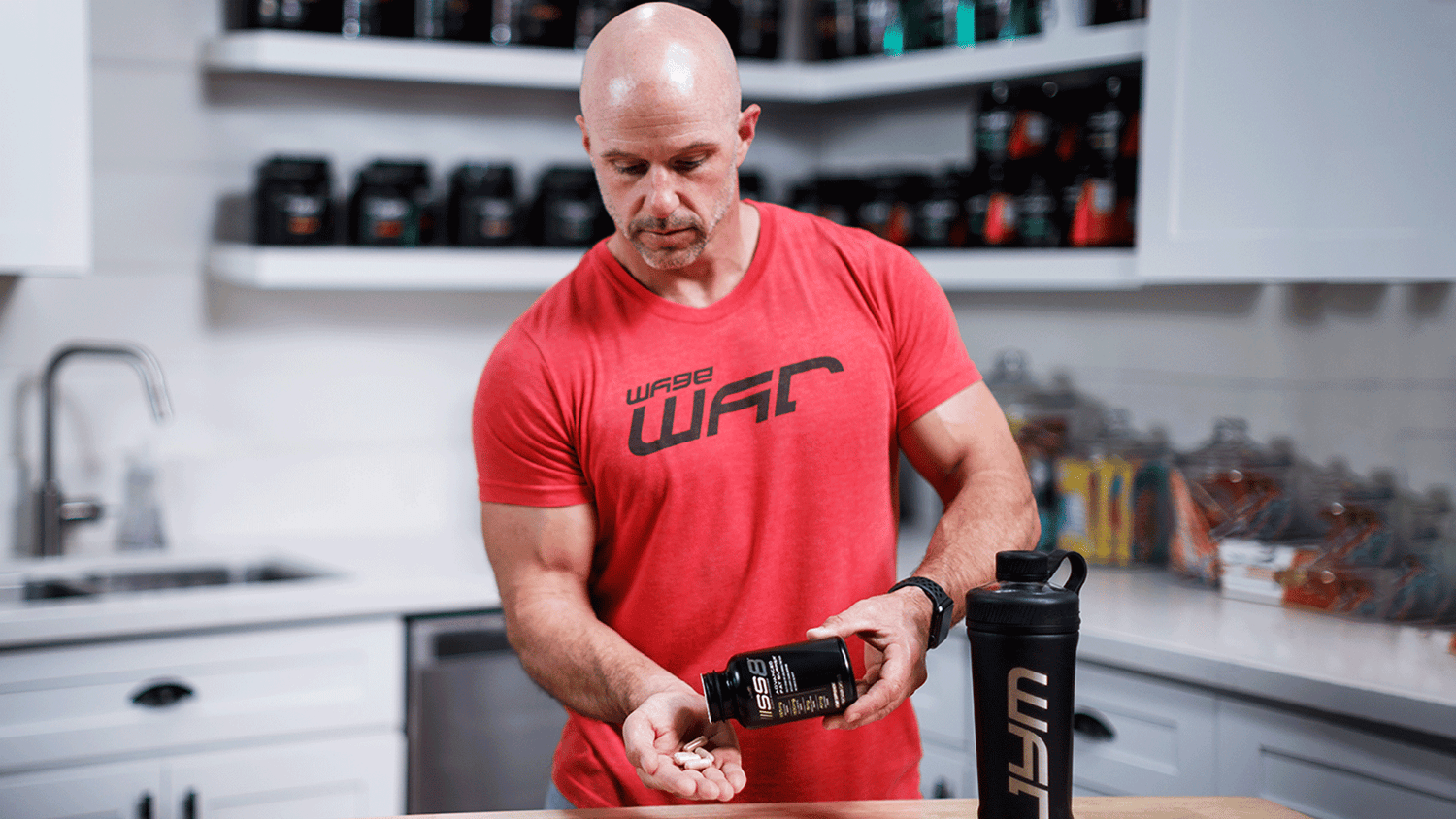Man in red Wage War shirt dispensing SS8 capsules into hand at kitchen counter, JYM shaker bottle and stocked supplement shelves.