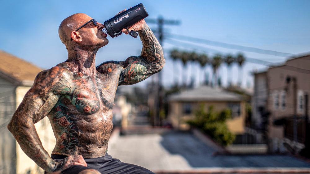 Jim Stoppani sips from a black JYM shaker on a sunny rooftop, tattoos across chest and arms - The Best Way to Use BCAAs.