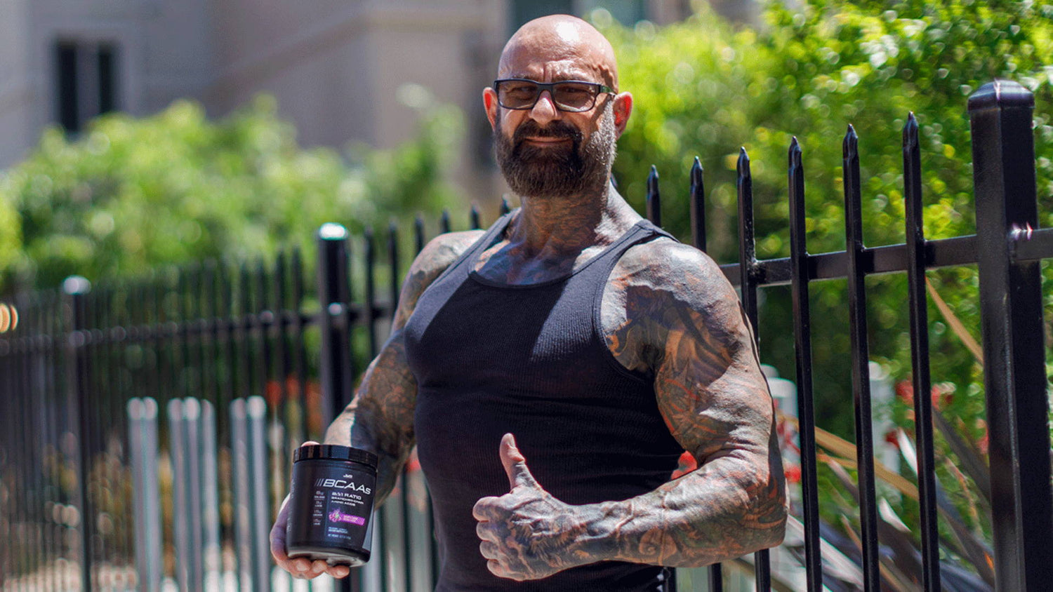 Muscular tattooed man in glasses and black tank top holds JYM BCAAs tub outside near greenery, Pre Workouts Must Have BCAAs.