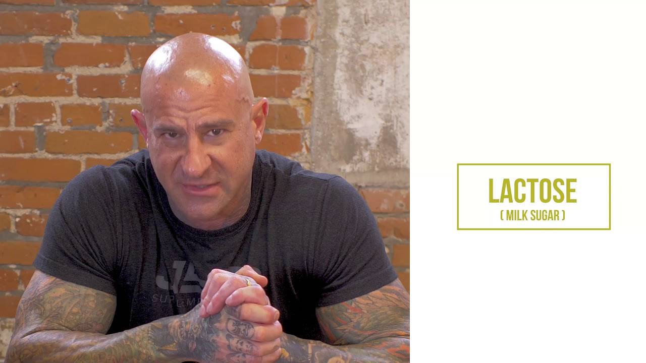 Jim Stoppani with tattooed arms crossed in front of brick wall, "Lactose (Milk Sugar)" label, on protein powder stomach issues.