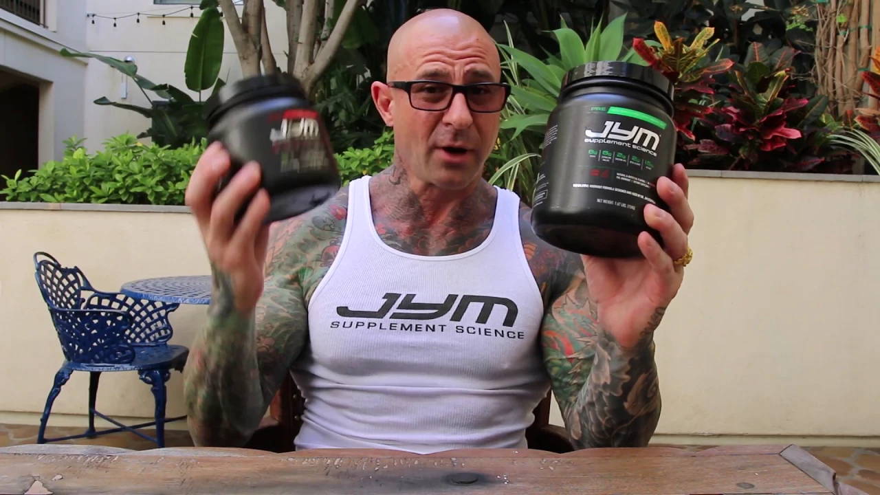 Bald, tattooed man in JYM tank top holds up two black supplement tubs on a plant-filled patio, discussing BCAAs pre and post workout.