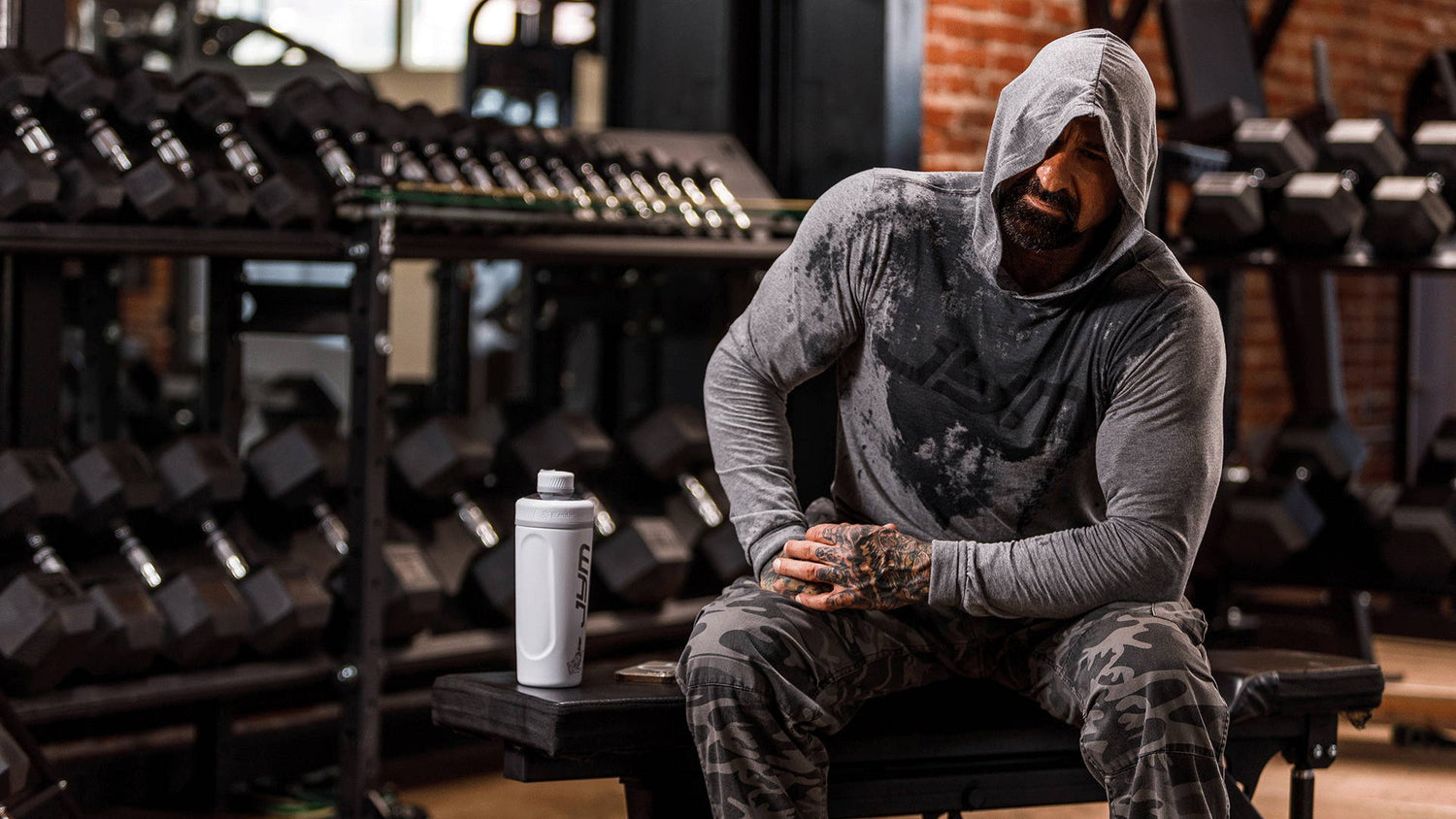 Bearded athlete in grey hood slumped on gym bench by white JYM shaker and dumbbell rack—How Real is the Risk of Overtraining.
