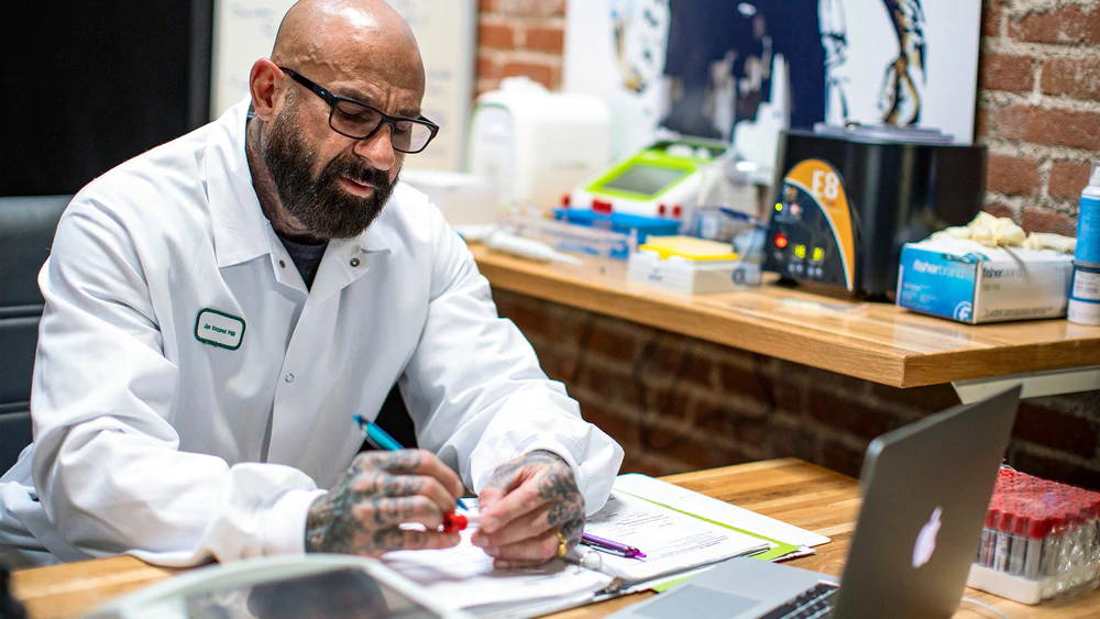 Dr. Jim Stoppani in lab coat and glasses writing notes beside Fisher Scientific supplies and E8 analyzer, Real Science vs. Broscience.
