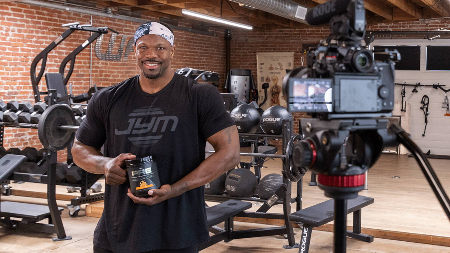 Michael Norwood in a black JYM shirt holds a Pre JYM X tub in a brick-walled gym while filming a testimonial on camera.