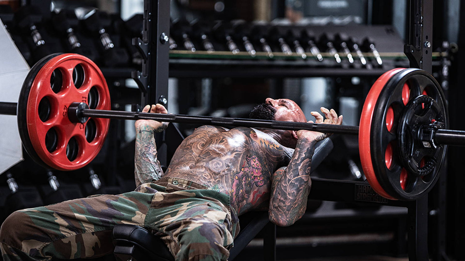 Bench Press Masterclass: shirtless tattooed lifter mid-rep on flat bench, barbell with red plates above chest, dumbbell rack behind.