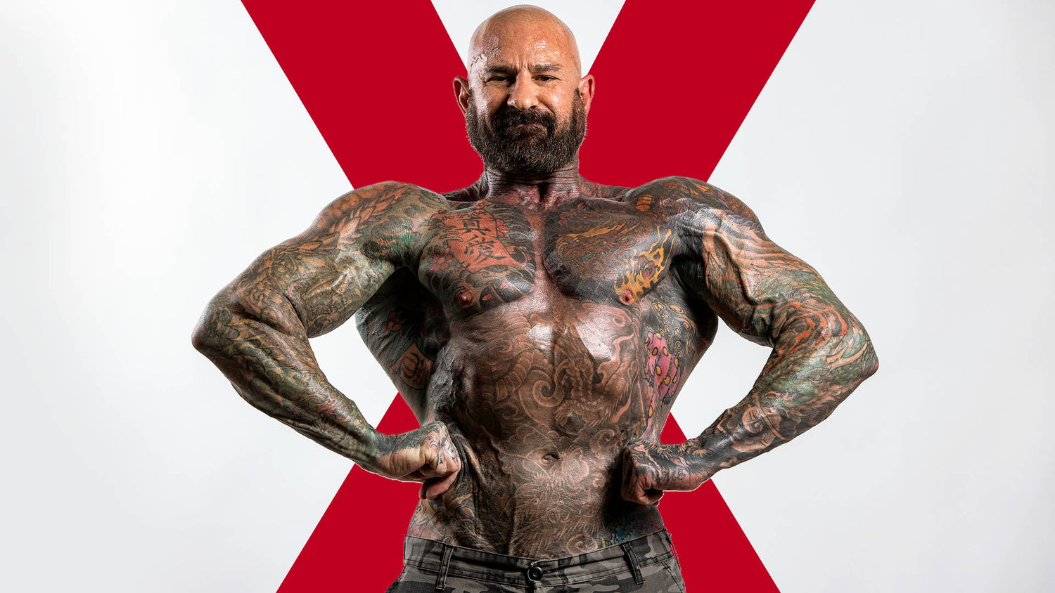 Tattooed Jim Stoppani standing shirtless in camo pants, flexing arms outward against a red X, for the X-Frame Physique article.