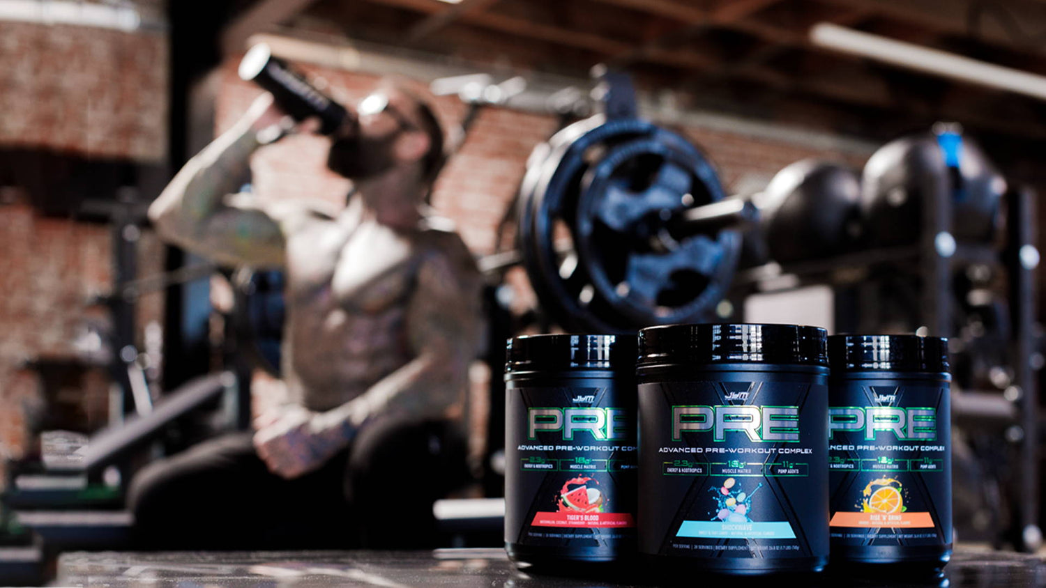 Three Pre JYM X tubs—Tiger's Blood, Shockwave, Rise N Grind—staged by a barbell as a tattooed lifter drinks from a shaker.