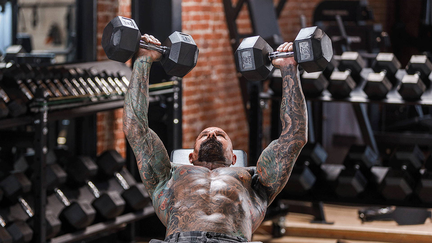 Tattooed lifter lying on bench pressing two black hex dumbbells overhead in a brick gym, for "Bigger Chest, Stronger Bench".